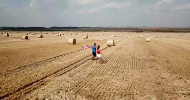 A young happy couple runs carefree and jumps on a wheat field. Holding the USA flag — Vídeo de Stock