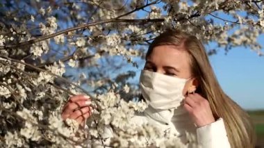 Beyaz maskeli sarışın kız çiçek açan meyve ağaçlarının arka planında selfie çekiyor.