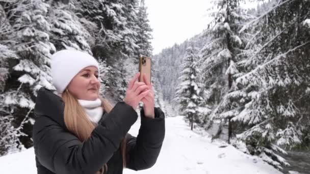 Chica rubia sostiene un teléfono inteligente en sus manos y toma fotos de un hermoso bosque de invierno — Vídeos de Stock