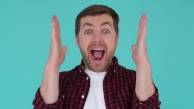 Close up portrait of young amazed guy shouting wow raising hands up, turquoise studio background
