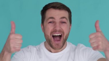 Young guy in a white T-shirt actively showing thumbs up on a turquoise background in the studio