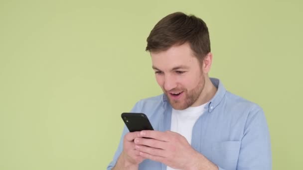 Portrait of happy young guy enjoying success on mobile phone in studio on olive background. — Stock Video