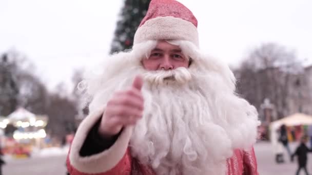Uśmiechnięty Święty Mikołaj podnoszący kciuk, super znak. Sanda na ulicy w pobliżu choinki — Wideo stockowe