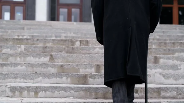 Um velho avô de casaco preto sobe as escadas, andando com força. O idoso de 80 anos — Fotografia de Stock