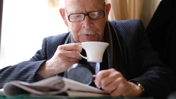 Gri saçlı büyükbaba bir kafede oturmuş çay içiyor ve gazete okuyor.. — Stok fotoğraf