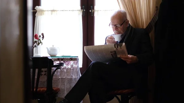 Avô de cabelos grisalhos está sentado em um café bebendo chá, ele está lendo um jornal. — Fotografia de Stock