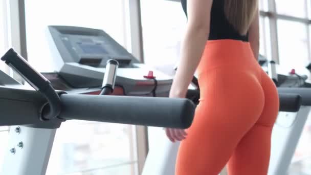 Chica atlética en el gimnasio, corre en una cinta de correr, vestida con pantalones de chándal naranja. — Vídeos de Stock