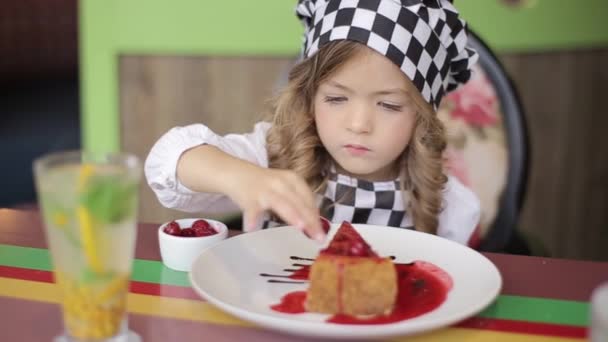 Забавная маленькая девочка прячется за белым столом и смотрит на вкусный торт — стоковое видео