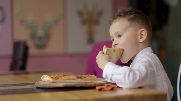 Der kleine Junge in der Küche isst eifrig eine kleine Pizza — Stockvideo