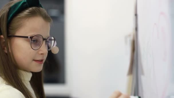 La niña dibuja un marcador en la pizarra de la escuela. Niño inteligente con gafas para mejorar la vista — Vídeos de Stock