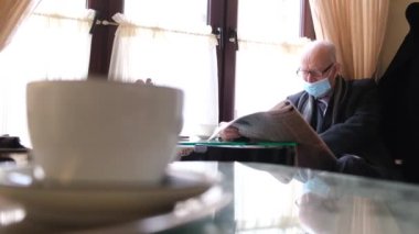 Gri saçlı, gözlüklü ve siyah ceketli bir adam kafede kitap okurken yeni bir gazete okuyor.