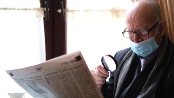 Anciano en gafas y una chaqueta negra está sentado en una silla junto a la ventana leyendo un periódico fresco — Vídeos de Stock