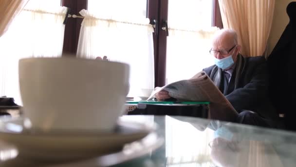 Un hombre de pelo gris con gafas y una chaqueta negra lee un periódico fresco mientras lee en un café — Vídeo de stock