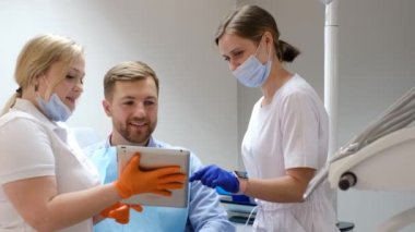 Diş hekiminde sandalyede oturan, maske takan genç bir adam diş tedavisi için tablet kullanıyor. Modern bir diş sağlığı kliniğinde diş tedavisi.