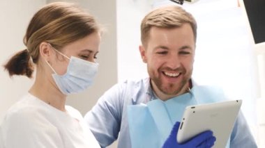 Kadın ortodontist diş muayenehanesinde genç bir adama tavsiye veriyor, elinde tablet olan bir doktora. Modern bir diş sağlığı kliniğinde diş tedavisi.
