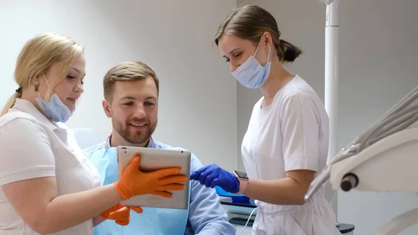 一个坐在牙医椅子上的年轻人 戴着口罩的医生们用一块石碑来建议如何修复牙齿 现代牙科诊所的牙科治疗 — 图库照片