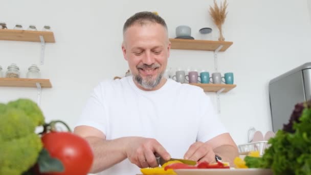 Χαμογελαστός Σεφ Κόβει Φρέσκα Λαχανικά Καθισμένος Στο Τραπέζι Της Κουζίνας — Αρχείο Βίντεο