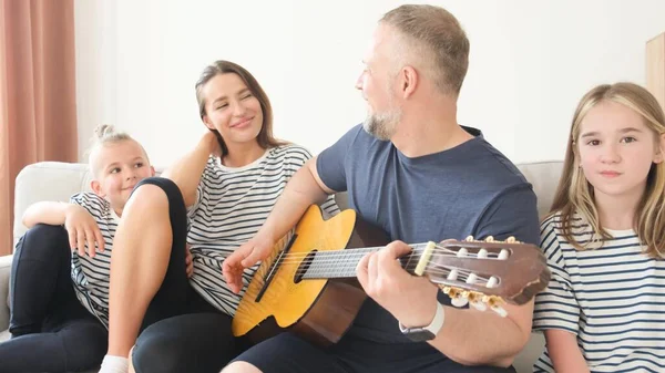 Mutlu bir aile kanepede oturup dinleniyor, babam karısı ve çocukları için gitar çalıyor. Gülümseyen genç baba gitar çalıyor, mutlu aile, mutlu anılar..