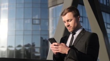 Akıllı telefon tutan ve gülen mutlu bir iş adamı. Elinde bir aletle neşeli bir adam kazandı. İş kıyafetleri içinde bir adam gökdelenin yanında duruyor ve haberleri okuyor.