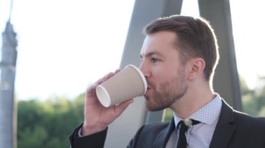 Güneş ışığı arka planında kahve içen genç iş adamının yakın portresi. Bir ofis çalışanının portresi, kameraya bakıyor ve gülüyor. İş konsepti