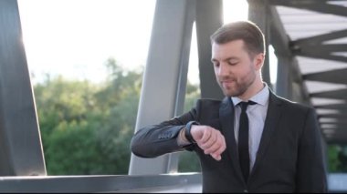 Takım elbiseli genç adam akıllı saate bakıyor. Sokaktaki işadamı müşterilerle görüşmek için bekliyor. Ağır çekim. Çerçevede yumuşak güneş ışığı