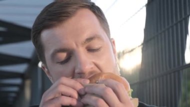 Yemek molasında iş adamı hamburger ya da sandviç yiyor, kapanıyor, iş merkezinin yanında duruyor. Şehirli hippi iş adamı, ağır çekim.