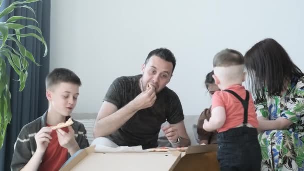 幸せな家族は自宅でソファに座ってピザを食べる 箱からおいしいイタリアのピザを食べている子供たちと夫と妻 ピザの宅配 — ストック動画