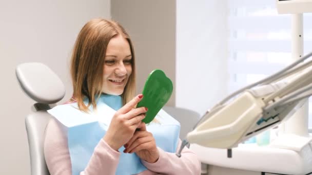 年轻的心满意足的微笑着的女人 带着完美的笑容 坐在牙科诊所里看着镜子 大姆指向上的女人 — 图库视频影像