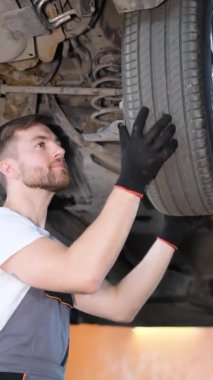 Dikey video, profesyonel bir araba tamircisi bir araba tamirhanesinde kaldırma makinesinin kralı altında çalışıyor. Araba servisi, elektrikli arabaların tamiri.