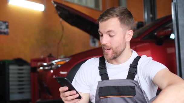 一位英俊的汽车修理工用智能手机 看着相机 开心地笑着 汽车服务的概念 机械师穿着制服使用小玩意的机械师 — 图库视频影像