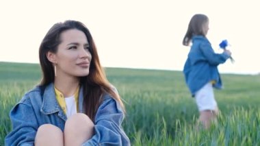 Anne ve kızı buğday tarlasında. Kadın oturuyor ve doğanın tadını çıkarıyor. Kızım elinde çiçeklerden bir buket çiçek tutuyor.