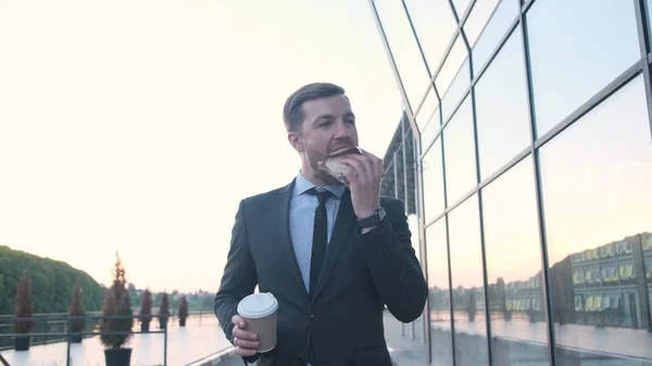 Young handsome businessman holding a cup of coffee, eating a burger on the street. Happy bearded man holding delicious food. Happy bearded man holding delicious food.