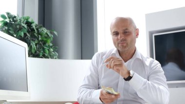 Başarılı bir iş adamı masaya bir tomar para atar ve kameraya bakar. Zengin işadamı para saçıyor, ofiste dinleniyor.