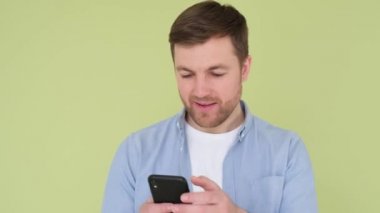 Elinde akıllı telefon olan genç Avrupalı adam sarı-yeşil stüdyo arka planında izole edilmiş aygıt ekranını işaret ediyor..