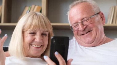 Mutlu son sınıf çifti, evdeki kanepede otururken akıllı telefondan selfie çektirirken eğleniyor. Modern aletler kullanarak yaşlı çiftlere gülüyorlar.