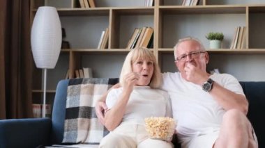 Happy elderly married couple watching TV sitting on sofa, elderly couple eating popcorn and laughing. Grandpa and grandma are happy together in retirement