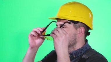 A man in a construction helmet he wears goggles on a background of chromakey. Portrait of a construction worker, he looks at the camera and laughs