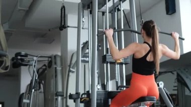 Atletik kadın spor salonunda sırt kaslarını çalıştırıyor, ana kas gruplarını pompalıyor. Kız kollarını simülatörde sallıyor. Spor salonundaki güzel beyaz kadın.