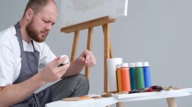 A professional artist mixes a palette of paints for painting. Behind him is an easel with a white canvas. Young male artist paints a picture in the studio