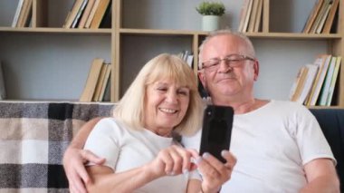 A happy elderly married couple is sitting in the living room and chatting online with their relatives and children. Modern means of communication
