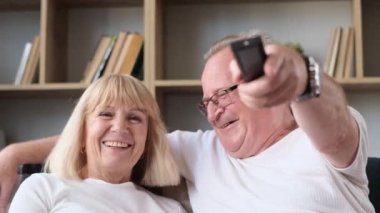 A happy elderly couple is sitting on the sofa and watching a movie. Time together. Joint family vacation.