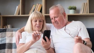 An elderly husband and wife are sitting on the sofa in the living room and mastering a smartphone. Elderly people and technology