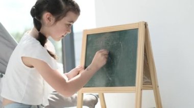 A cute little girl with black pigtails draws with chalk and felt-tip pens on an easel. Childrens creativity. Happy childhood.