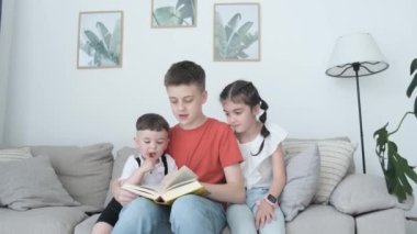 Three wonderful, cute children are sitting on the couch in the living room and looking at a book. Family time together. The concept of a happy childhood.
