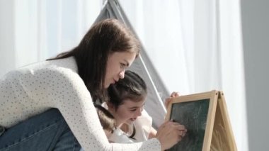 A young mother draws with chalk on an easel together with her little son and daughter. Mothers play with children. Happy childhood