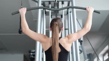 Atletik kadın spor salonunda sırt kaslarını çalıştırıyor, ana kas gruplarını pompalıyor. Kız kollarını simülatörde sallıyor. Spor salonundaki güzel beyaz kadın.