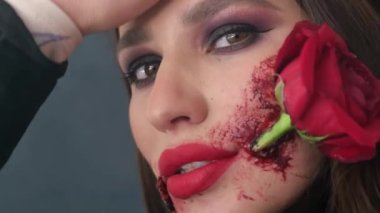 Glamorous brunette woman with makeup for Halloween, rose flower in her mouth. Portrait of a girl holding a red rose in her mouth. High quality 4k video