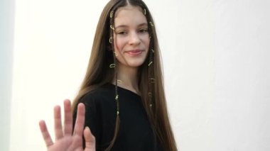 European teen girl waves her hand at the camera, she is on a white background. Beautiful child of 12 years smiling, long straight hair