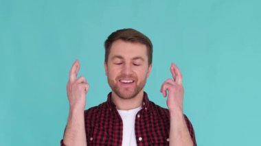 Concentrated american young man holding fingers crossed and making wishes isolated on turquoise background. He is wearing a red shirt and a white T-shirt
