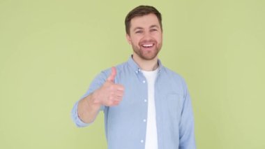 Young handsome man with beard in denim shirt on yellow-green background, sign of success making positive hand gesture, smiling and happy thumb up. Cheerful facial expression